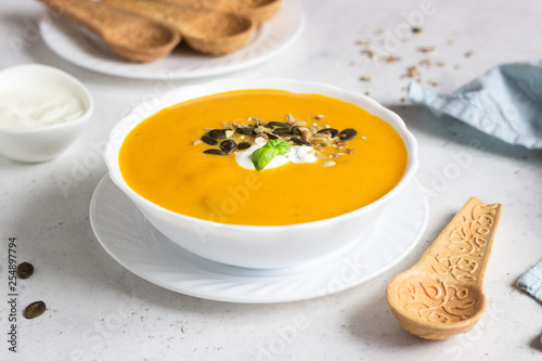 Fresh homemade pumpkin creamy soup served with sour cream, basil and seeds in a white bowl on a light grey concrete background. View from above, flat lay.