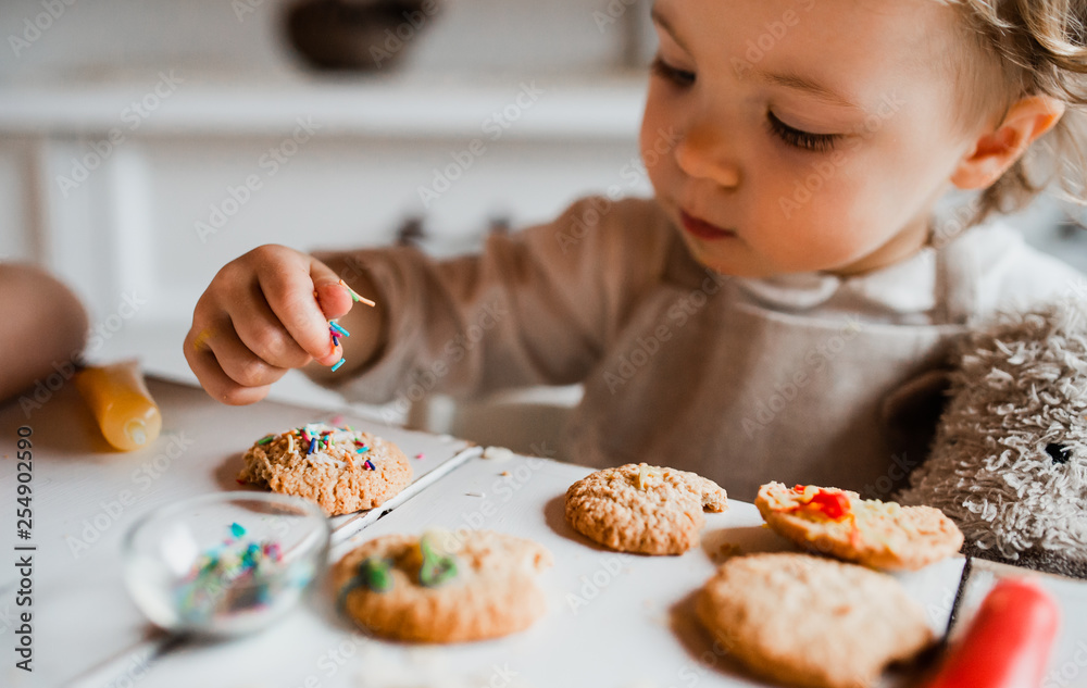 A small toddler girl sitting at the table, decorating cakes at home. A small toddler girl sitting at the table, decorating cakes at home.