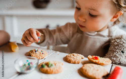 A small toddler girl sitting at the table, decorating cakes at home.