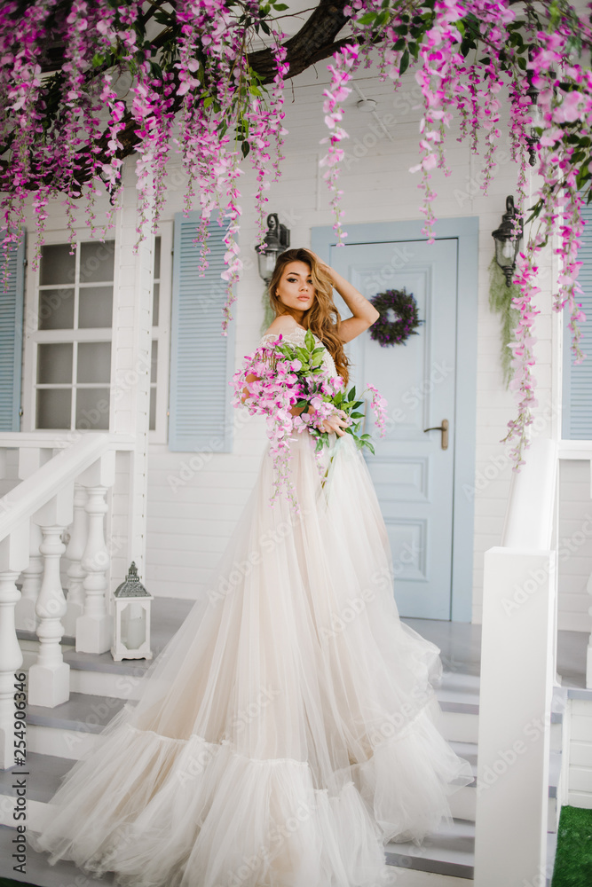 Charming girl in a long white dress standing on the stairs with a bouquet in her hands