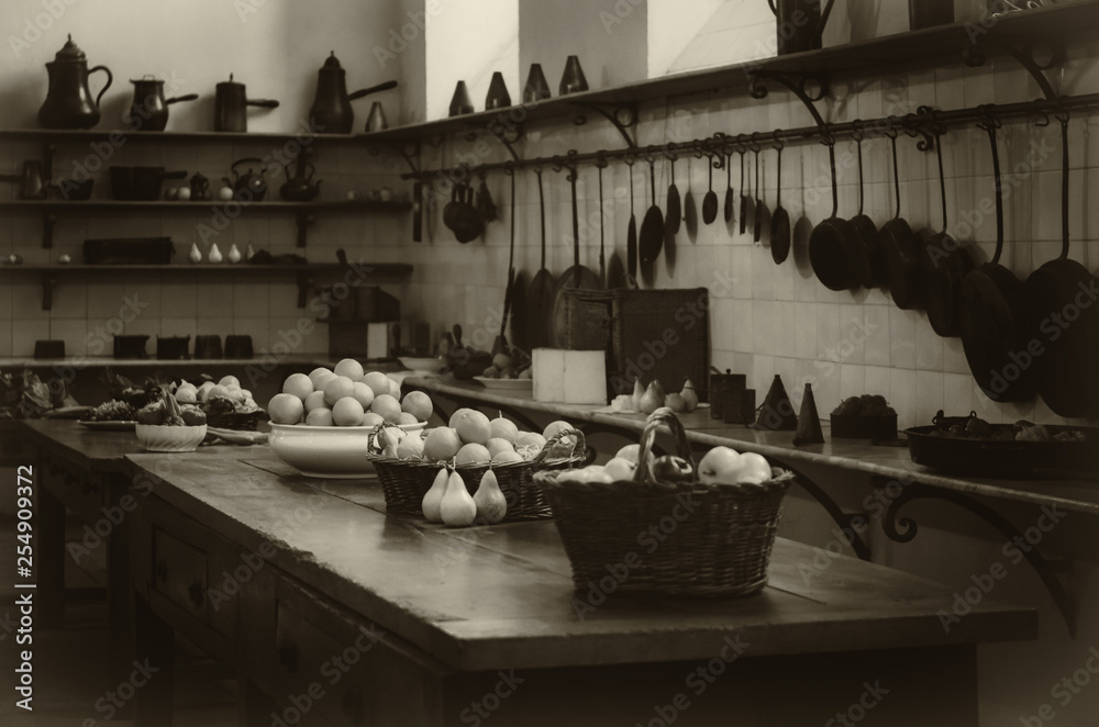 vintage looking image of an antique XIX century old kitchen with tools, pans, pots and food ingredients all over che benches and tables