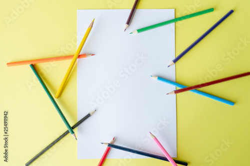 A rainbow of colored pencils are scattered randomly on a white vertical sheet of paper that is lying on a yellow background. Top view