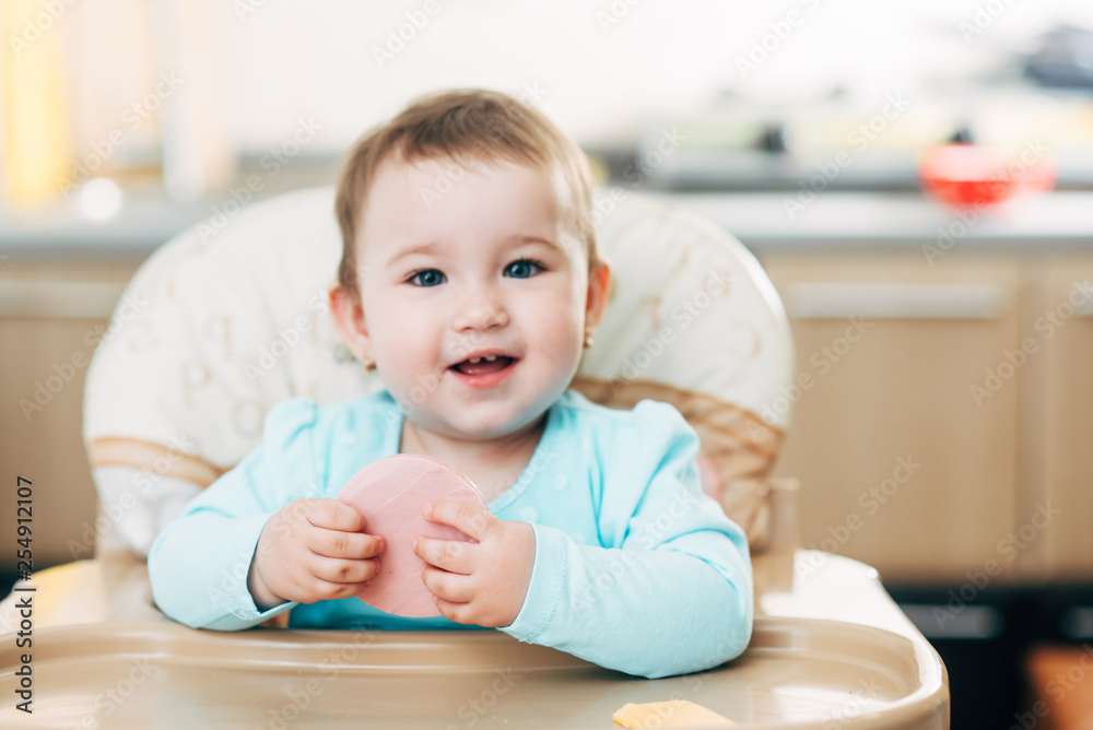 a little girl in a high chair with a piece of boiled sausage in her hands a little girl in a high chair with a piece of boiled sausage in her hands