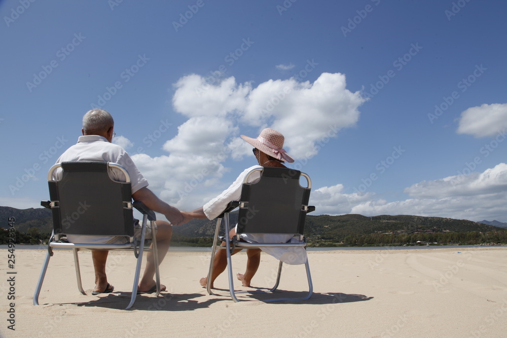 coppia di anziani di spalle si tiene la mano seduti in spiaggia