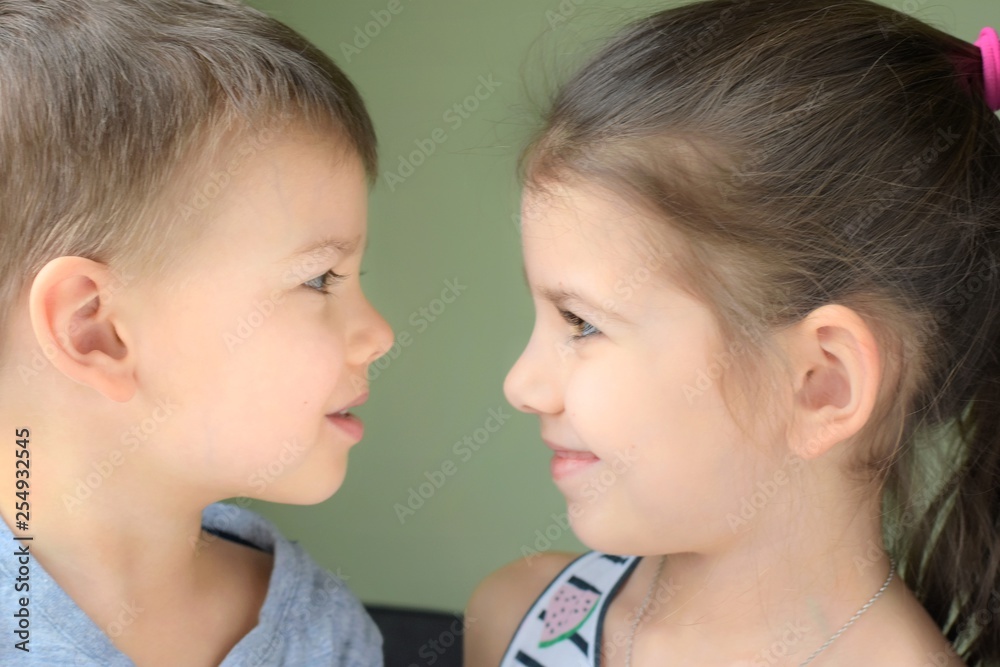 White caucasian toddler boy and girl look at each other with love on blurred neutral background. Cute little brother and beautiful sister with happiness faces looking at each other and smiling 