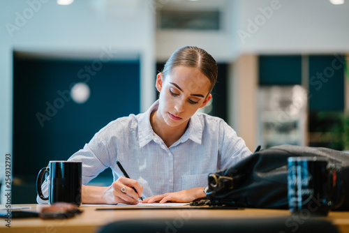 A pretty Caucasian lady writing on her notebook inside a cafe. She is wearing a neat tops while enjoying her coffee.