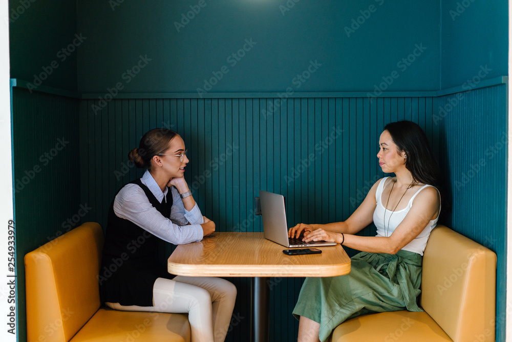 Two delightful women talking about an archive inside a bistro. They are taking a gander at their work in a PC while sitting on a dark colored lounge chair. Two delightful women talking about an archive inside a bistro. They are taking a gander at their work in a PC while sitting on a dark colored lounge chair.