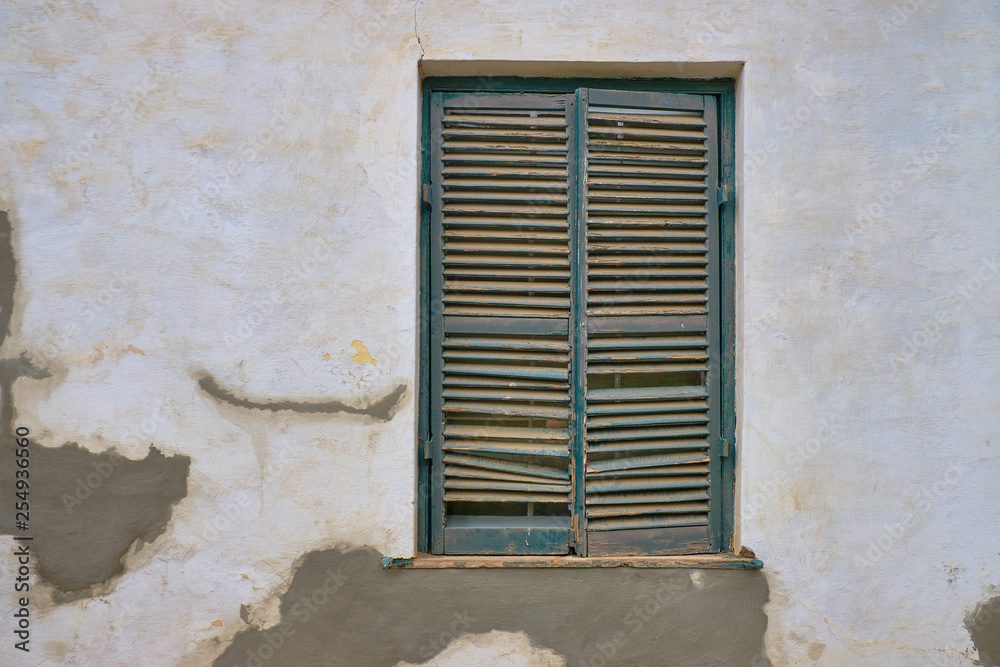 Minimalist capture of a single rural window on a white textured wall with plaster repairs.  Window has dark grey shutters in a state of needing repairs.