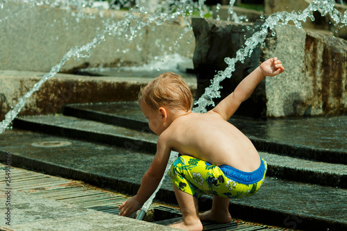 Funny baby boy trying to cauch water stream in fountain. Cute toddler playing in the city fountain