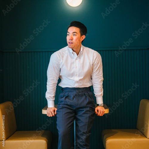 Portrait of a young, gorgeous hunk Chinese Asian business man in a white long sleeves and black pants. He looks deliberate as he leans against a table in the office pantry during the day. 