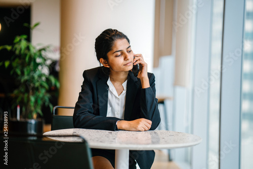 A young Indian businesswoman is sitting inside a cafeteria while talking to a friend over the phone seriously. 