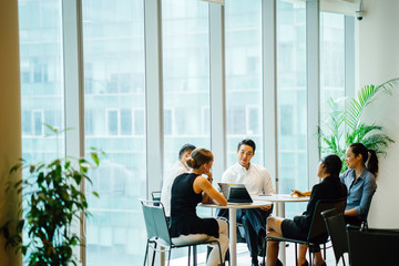 A diverse team sits around a table in a meeting room to have a business meeting to discuss plans. The group is international with Asian and white team members and they are all professionally dressed.