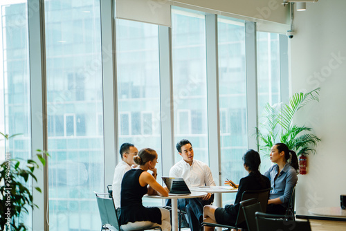 A diverse team sits around a table in a meeting room to have a business meeting to discuss plans. The group is international with Asian and white team members and they are all professionally dressed.