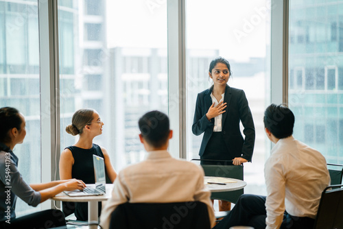 An Asian Indian businesswoman is ingeniously explaining with her diverse team in a conference room. She's wearing a black blazer and white blouse with a black skirt. photo