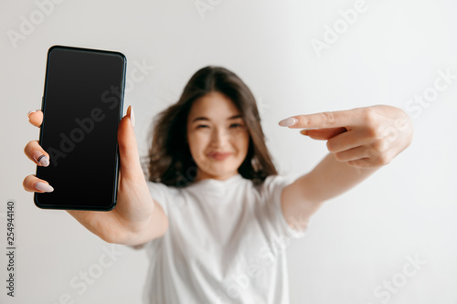 Portrait of a confident casual asian girl showing blank screen of mobile phone isolated over gray background at studio.