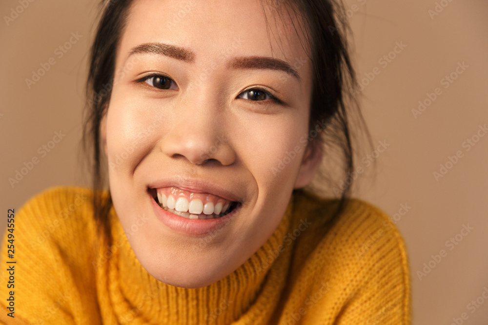 Image closeup of joyous asian woman wearing sweater smiling while posing at camera in studio Image closeup of joyous asian woman wearing sweater smiling while posing at camera in studio