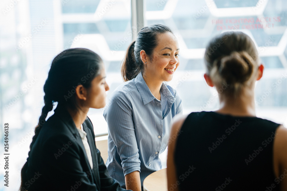 Image of a young Asian Chinese manager chatting in their office with her team. As she listens to her colleague speaking about their new project, she smiles back. Image of a young Asian Chinese manager chatting in their office with her team. As she listens to her colleague speaking about their new project, she smiles back.