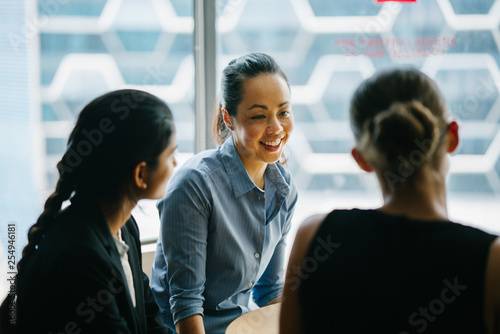 Image of a young Asian Chinese woman manager in their office chatting with her team. She is smiling as she listens to her colleague discussing something about their new project.
