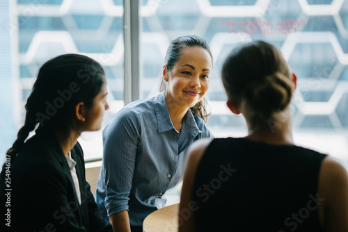 Image of a young Chinese Asian woman manager chatting with her team in their office. She smiles back as she listens to her colleague talking about their new project.