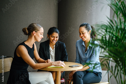 A diverse group of three young women sit around a table and smile while having a casual discussion. They are professionally dressed and are in a meeting room in an office.