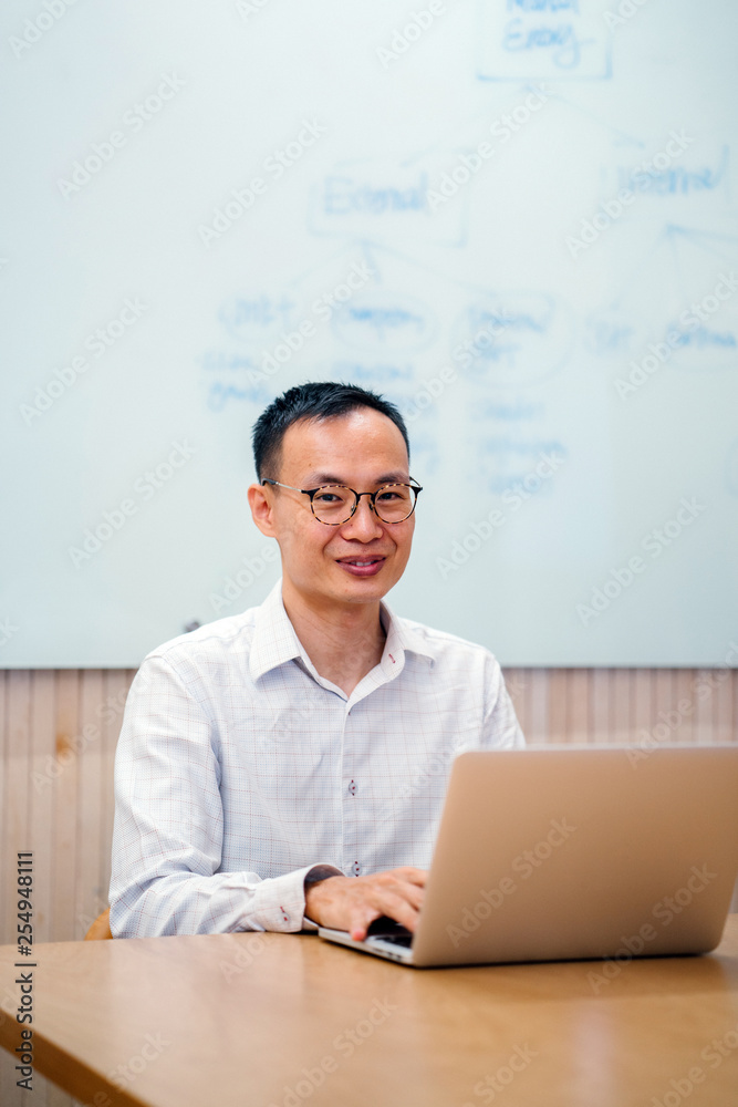 A middle-aged Asian businessman from China is working on his laptop computer while in a meeting room in his office. As he looks up from his work, he smiles. A middle-aged Asian businessman from China is working on his laptop computer while in a meeting room in his office. As he looks up from his work, he smiles.