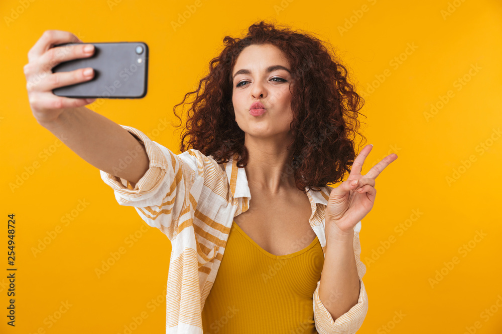 Image of positive woman 20s with curly hair smiling and taking selfie photo on smartphone - obrazy, fototapety, plakaty 