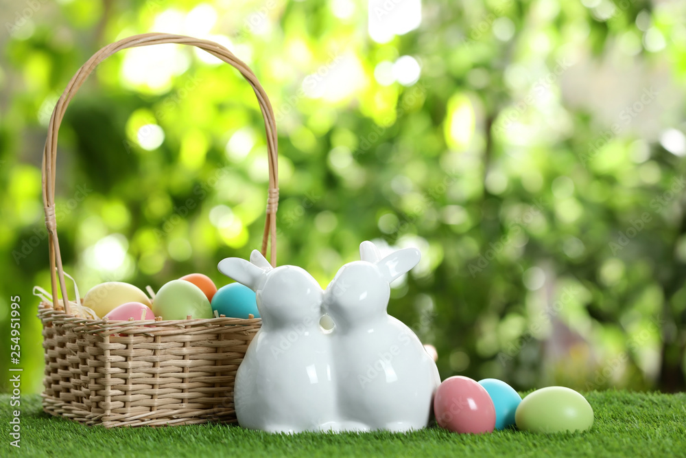 Cute ceramic Easter bunnies with wicker basket and dyed eggs on green grass against blurred background, space for text Cute ceramic Easter bunnies with wicker basket and dyed eggs on green grass against blurred background, space for text