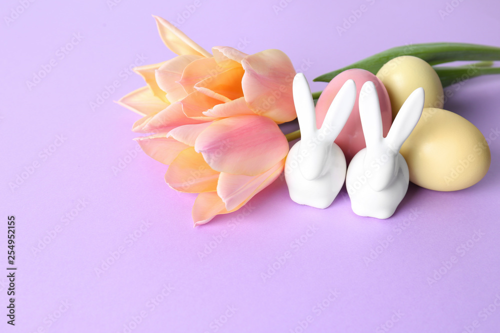 Cute ceramic Easter bunnies, dyed eggs and spring flowers on color background