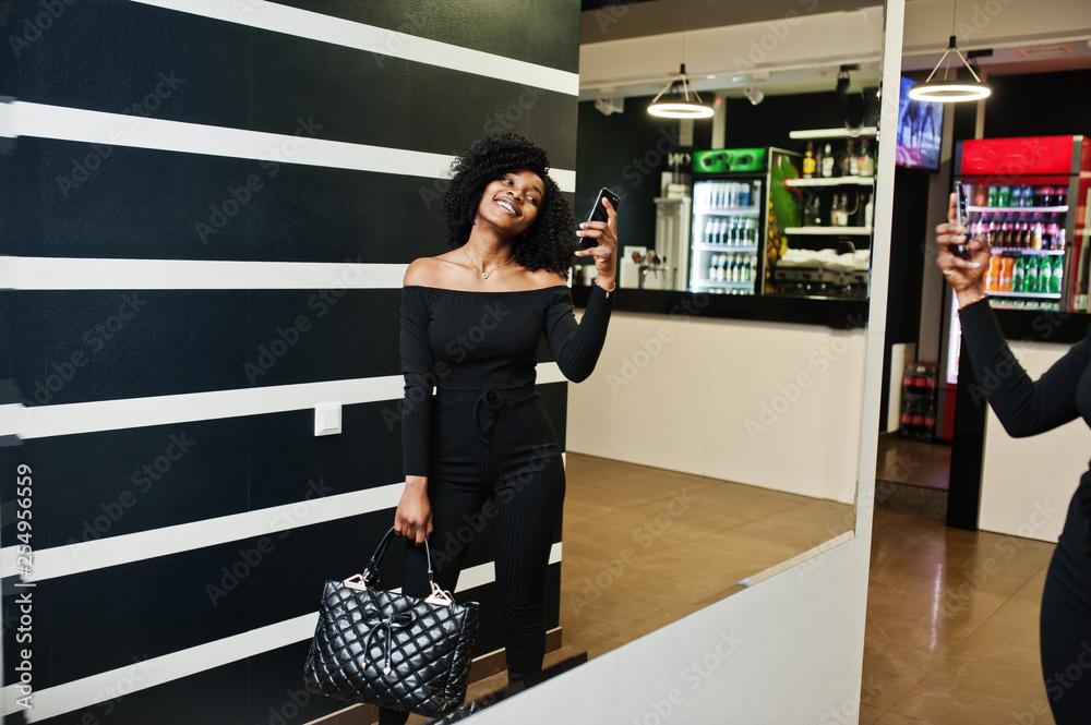 Fototapeta premium Fashionable young beautiful african american woman with afro hairstyle and hangbag posing wear in elegant black. Making photo at mirror by phone.