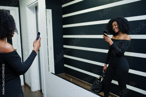 Fashionable young beautiful african american woman with afro hairstyle and hangbag posing wear in elegant black. Making photo at mirror by phone. photo