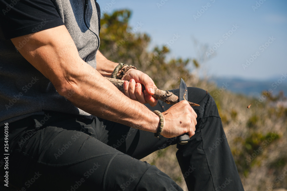 Man sitting on a rock in the nature carving a wooden stick - obrazy, fototapety, plakaty 