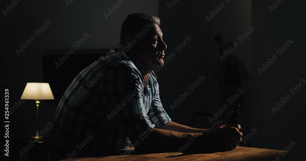Sad and thoughtful senior man alone in the dark with a bottle of beer in hands. Alcohol can not solve problems