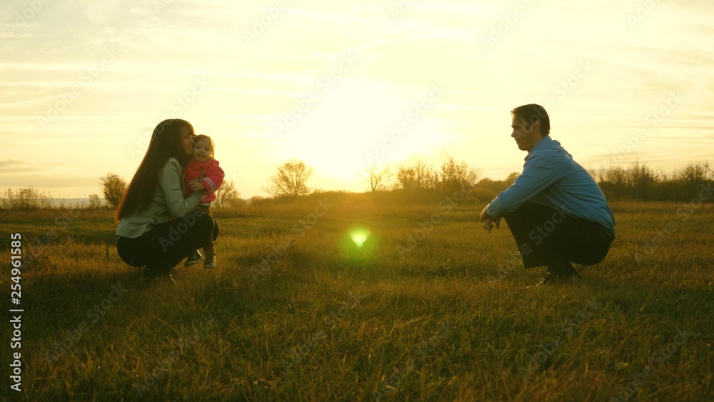 mom and dad play with kid on the grass at sunset. family happiness concept. baby goes on lawn from dad to mom. child takes first steps in park.