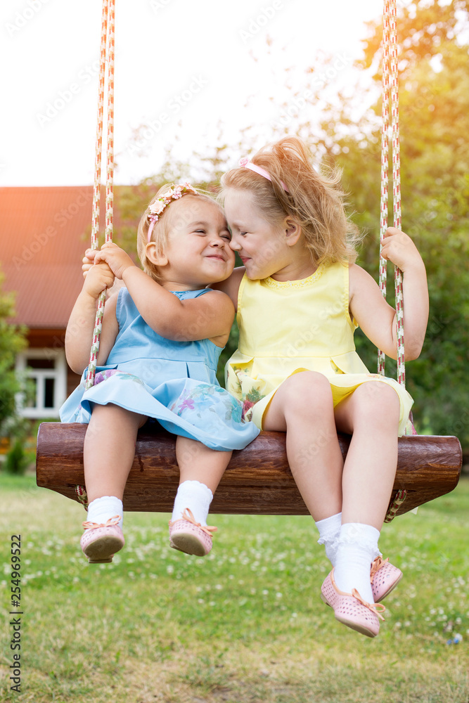 Two cheerful little caucasian blondie girls outdoors on swing in countryside Two cheerful little caucasian blondie girls outdoors on swing in countryside