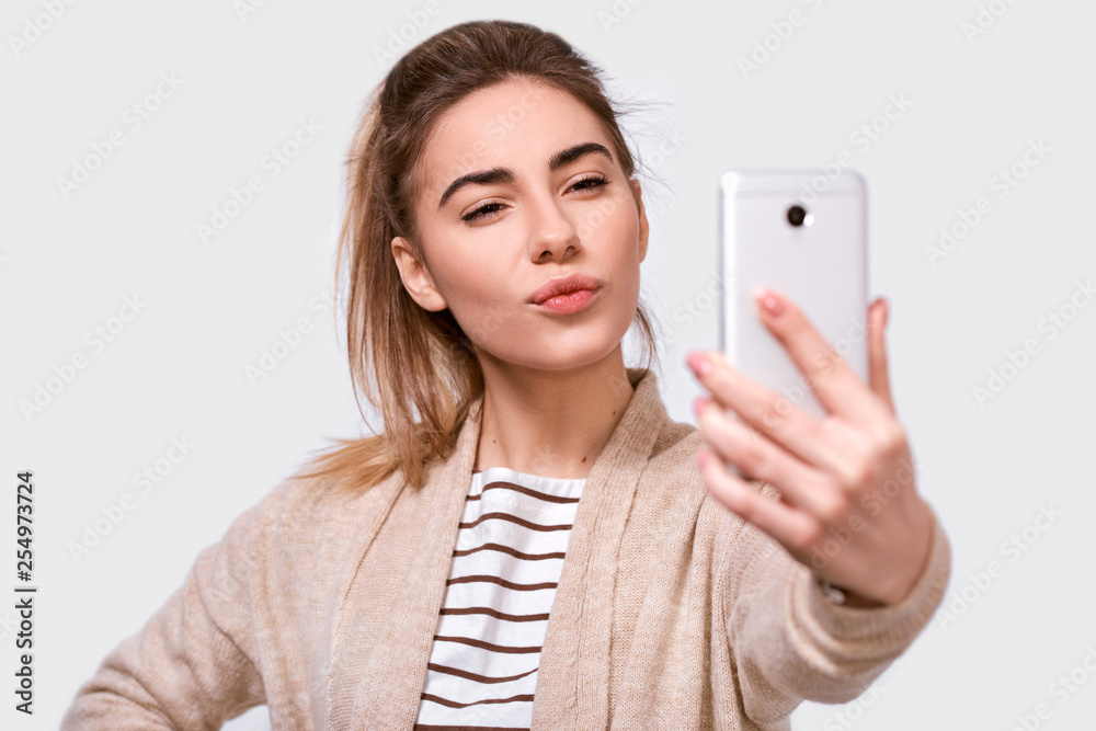 Studio close up studio shot of a pretty young European woman in casual clothes sending air kiss to his boyfriend while standing and taking a selfie isolated over white studio background. Studio close up studio shot of a pretty young European woman in casual clothes sending air kiss to his boyfriend while standing and taking a selfie isolated over white studio background.