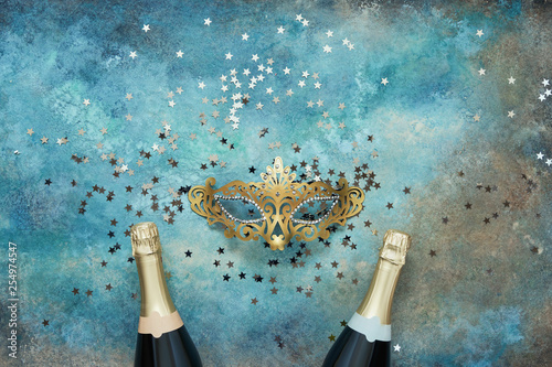 Two champagne bottles, golden carnival mask and confetti stars on blue background. Christmas background, top view, copy space