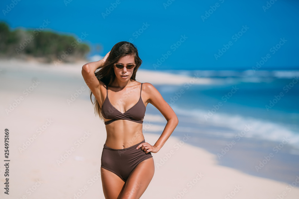 Beautiful tanned girl posing on beach with white sand and blue ocean.