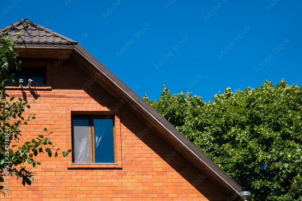 Detail of red brick house with window standing among lush green trees against bright blue sky