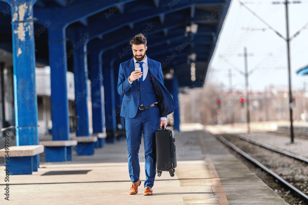 Full length of bearded businessman in formal wear holding luggage and using smart phone while walking at train station. Business trip concept. Full length of bearded businessman in formal wear holding luggage and using smart phone while walking at train station. Business trip concept.