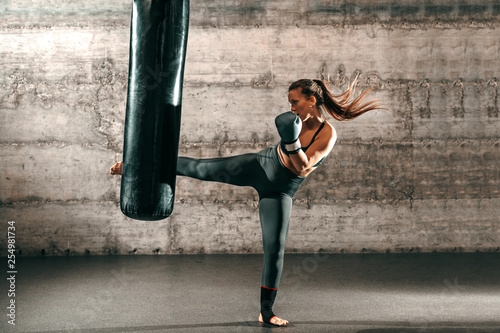 Photo Dedicated strong brunette with ponytail, in sportswear, bare foot and with boxing gloves kicking sack in gym