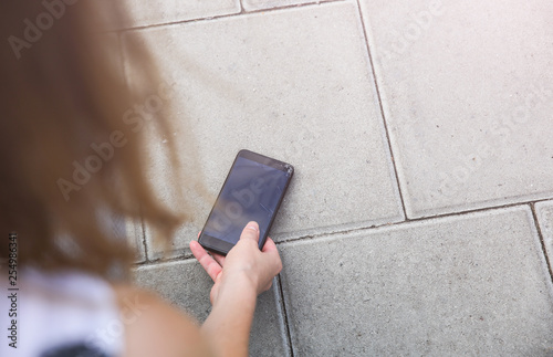 Woman is picking up broken smartphone lying on the road outdoors. Crashed screen from the accidental fall. Fragile modern technologies.