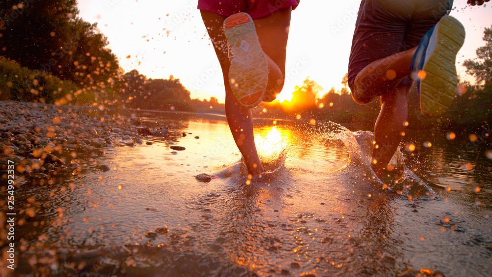 CLOSE UP: Glassy drops of water fly as athletic couple jogs in shallow river. - obrazy, fototapety, plakaty 