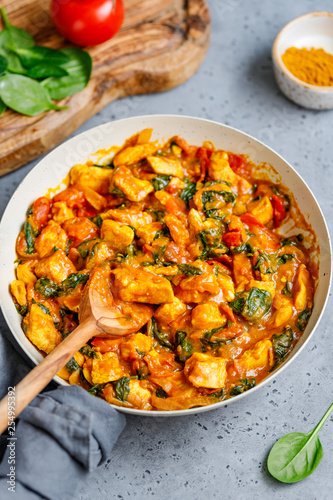 Bombay chicken curry with spinach, tomato and onion in a white frying pan on a kitchen table. Indian traditional cuisine.