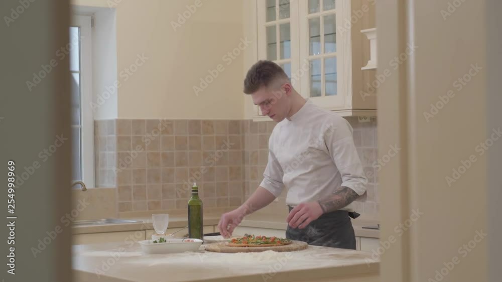 Professional chef in a uniform makes pizza in the kitchen at home. The chef sorts pizza with grated parmesan cheese. Size view. Cooking concept. Professional chef in a uniform makes pizza in the kitchen at home. The chef sorts pizza with grated parmesan cheese. Size view. Cooking concept.