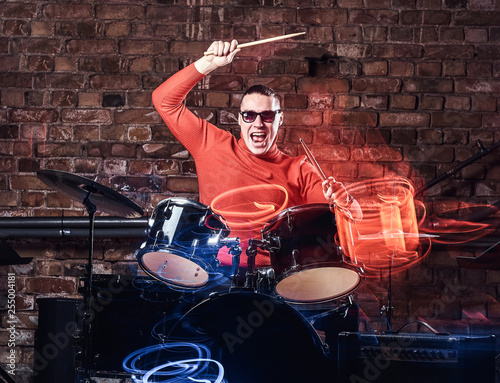 Young Stylish musician in sunglasses emotionally playing drums against brick wall background, perform in a night club. Photo with lighting motion effect