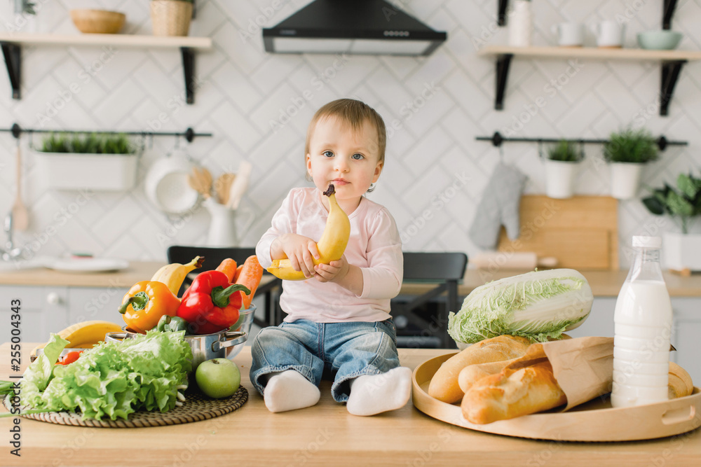 Beautiful little girl one year old, sitting on a table in the kitchen among fruits and vegetables and holding a banana in her hand. Modern kitchen on the background Beautiful little girl one year old, sitting on a table in the kitchen among fruits and vegetables and holding a banana in her hand. Modern kitchen on the background