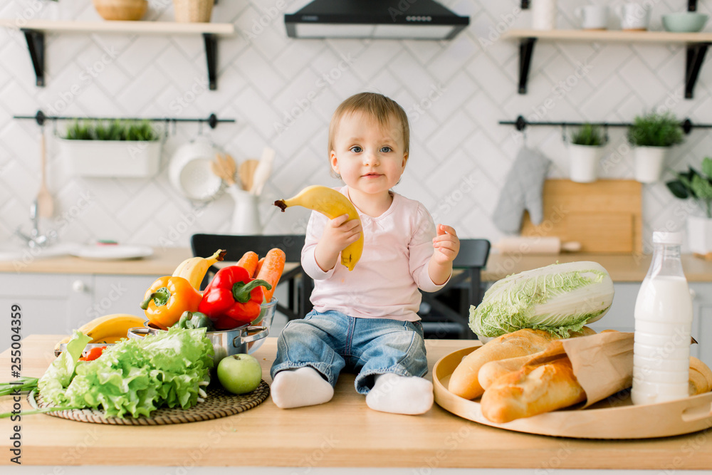 Beautiful little girl one year old, sitting on a table in the kitchen among fruits and vegetables and holding a banana in her hand. Modern kitchen on the background Beautiful little girl one year old, sitting on a table in the kitchen among fruits and vegetables and holding a banana in her hand. Modern kitchen on the background