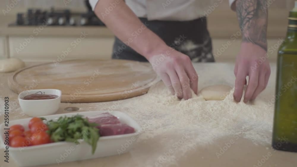 Close up hands of young pizza maker in cook uniform throws dough ball in the kitchen. Pizza ingredients lying on the table in the foreground. Food preparation. Camera moves right. Slow motion Close up hands of young pizza maker in cook uniform throws dough ball in the kitchen. Pizza ingredients lying on the table in the foreground. Food preparation. Camera moves right. Slow motion