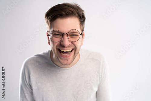 Portrait of young Caucasian guy enjoys funny story or joke, laughs joyfully, has toothy smile, wears white t shirt in one tone with background. Happiness and youth concept. © opolja
