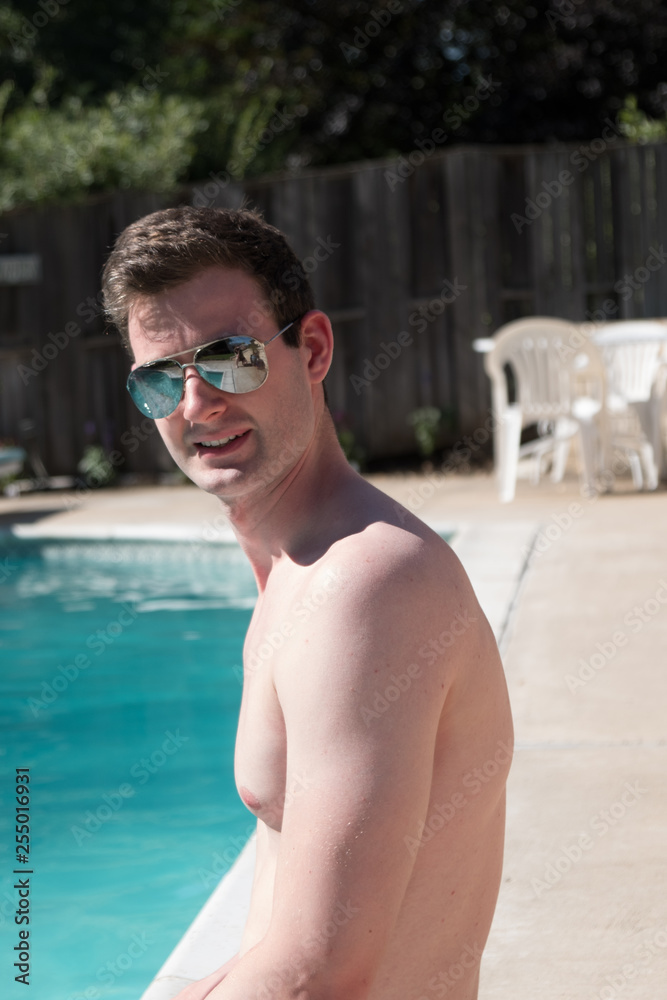 White man sitting on the edge of an outdoor swimming pool wearing aviator sunglasses head turned looking at the camera. Man sitting poolside by outdoor pool.
